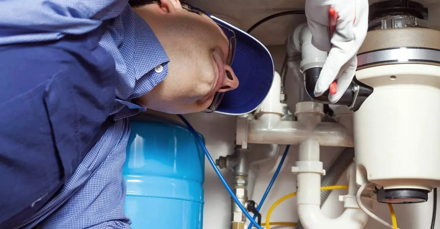 Gasfitero con gorra y guantes revisando tuberías bajo un lavadero, junto a un tanque azul de presión